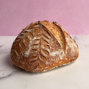 A sourdough boule with a golden crust featuring a hand cut leaf design, on a granite counter in front of a pink background. Dover Dollhouse Sourdough in Dover NH
